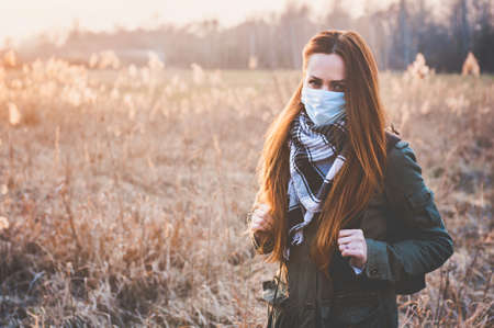 A girl in a field with a medical mask on her face, against the backdrop of a sunset. Concept on the theme of quarantineの写真素材