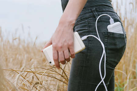 Power bank in the back pocket of a girl in a yellow field charges the phone, against the background of ears of wheat and sky, side view of a young beautiful woman.の写真素材