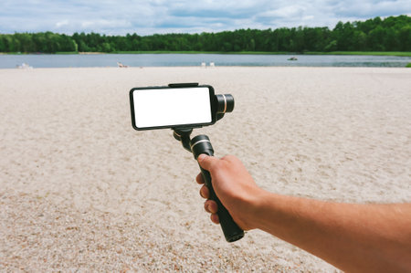 Mock up of a smartphone with a camera stabilizer in a man's hand. Against the backdrop of a sand beach and nature with a lakeの写真素材