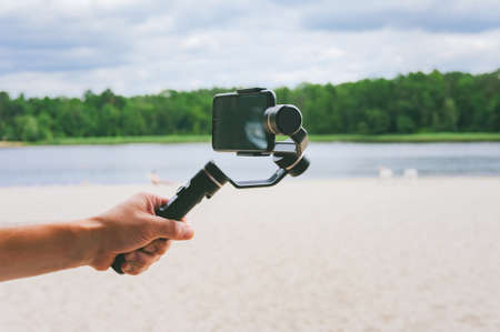 Smartphone camera stabilizer in a man's hand. Against the backdrop of a sand beach and nature with a lakeの写真素材