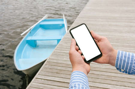 Mockup of the phone in the hands of a man Against the background of a boat on the water at a wooden pierの写真素材