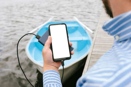 Mock-up of a smartphone with charging from Power bank in a man's hand. Against the background of a boat on the water at the wooden pierの写真素材