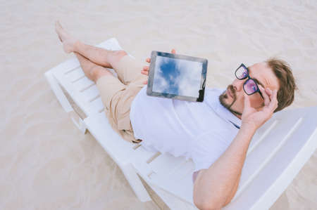 Mock up tablet in the hands of a man with glasses looking at the camera and lying on the beach on a...