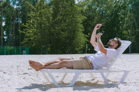 The guy lies on a sun lounger, takes a selfie on his smartphone and charges from the Power Bank. against the backdrop of the beach and forestの写真素材