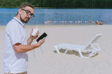 Portrait of a man with glasses charging a smartphone from a Power Bank on the beach. Against the backdrop of sand, sun lounger and waterの写真素材