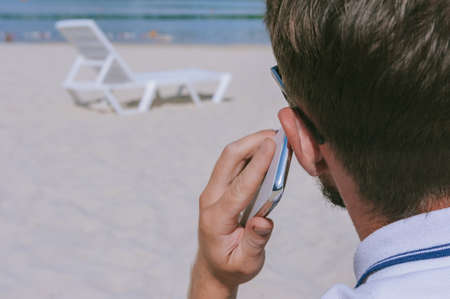 A man is talking on the phone on the beach. Against the background of sand, a chaise lounge from the waterの写真素材