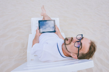 A mock-up of a black tablet in the hands of a man with glasses, lying on a beach chair. Against the background of sandの写真素材