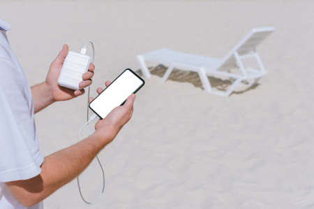 A man charges a smartphone from a Power Bank on the beach. Against the backdrop of sand and a white...