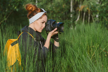 A girl in a gray jacket with a yellow backpack holds a professional photo-video camera in her hands. Against the background of green nature and forestの写真素材