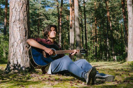 The guy plays the guitar sitting near the tree. Against the background of the forestの写真素材