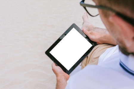 Mock-up of a tablet in the hands of a man lying on a beach on a sun lounger. Against the background of sandの写真素材