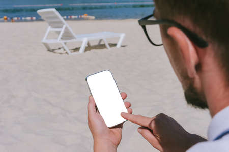 Finger touch, mock up smartphone in the hand of a man on the beach with a deck chair. Against the background of sand and waterの写真素材