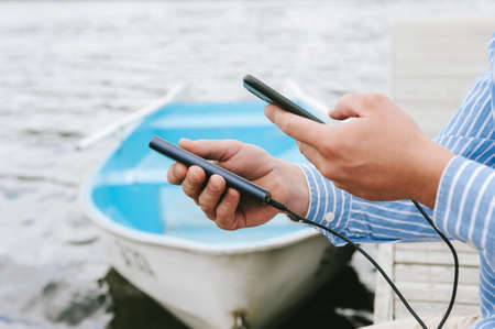 Close-up, Power Bank charging a smartphone in a man's hand. Against the backdrop of boats on the...