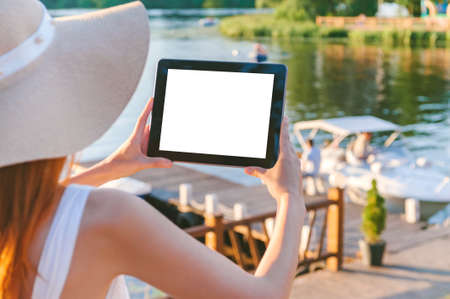 Mock up tablet in the hands of a girl in a big hat. Against the background of a wooden embankment with water and a boat. Over the shoulder viewの写真素材