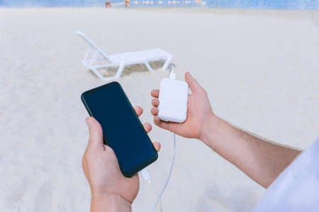 Power Bank Energy charges the phone into the hands of a man on the beach. Against the backdrop of sand and a sun loungerの写真素材