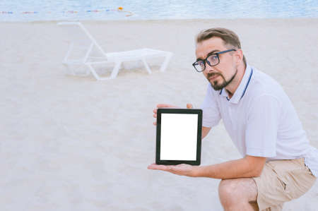 A man in glasses sits on the beach with a mock-up of a tablet in his hands. Against the backdrop of...