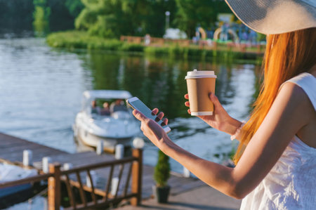 A girl with a hat holds a smartphone with coffee in her hands. In the background water embankments with a pleasure boatの写真素材