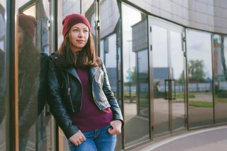 A beautiful girl in a leather jacket and a hat stands against the background of a glass buildingの写真素材