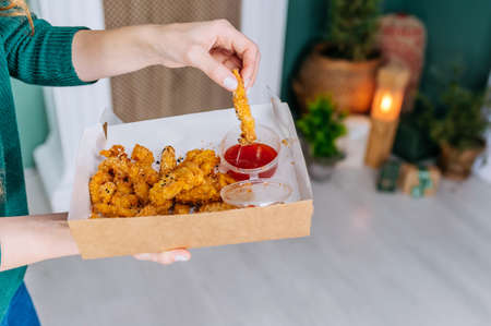 Close-up girl dips nuggets in sauce. On the background of the fireplace in the home. Delicious takeaway fast foodの写真素材