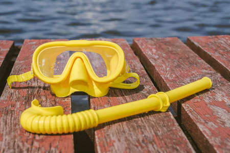 Close-up, yellow waterproof mask for snorkeling, diving with a snorkel on a wooden pier. Seascape background with blue water of sea or ocean, river, lakeの写真素材