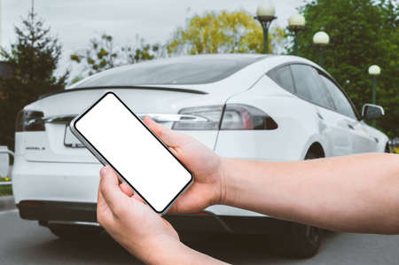 Close-up, Mockup of a smartphone in the hands of a man. Against the background of a white electric car in the cityの写真素材