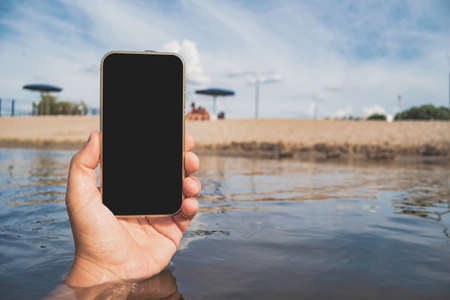 Phone in a man's hand. Against the backdrop of water, beach and blue skyの写真素材