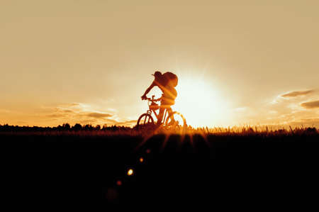 Silhouette of a young man, a woman cyclist on a sunset sky with clouds and sun. Against the backdrop of a yellow sunset skyの写真素材
