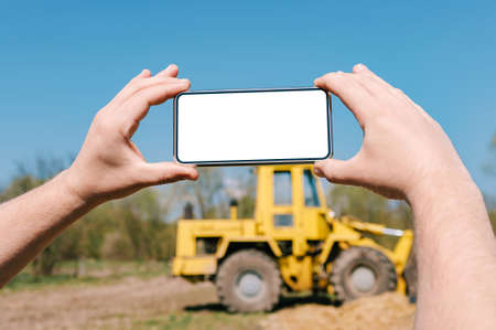 Smartphone mockup in mans hands. Against the background of a yellow excavator with a wheel loader in...