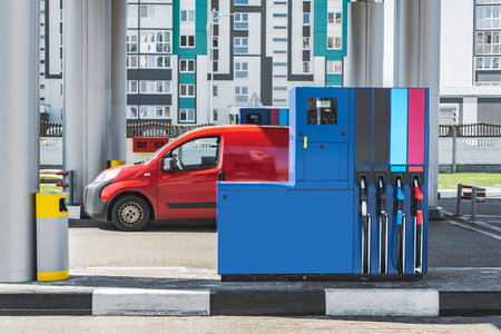 A blue gas pump with fuel pistols against the background of a red car standing at a gas station in a city with residential buildingsの写真素材
