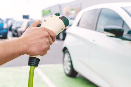 car charger in the hands of a man against the background of a power station for charging cars in an open parking lotの写真素材
