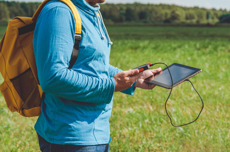 Close-up: a young man in a blue sweater with a yellow backpack on his back, holds a power bank in his hands and charges a digital tablet. Background of a green field with grassの写真素材