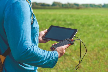 A power bank charges a digital tablet in the hands of a man. Nature background with blue sky and...