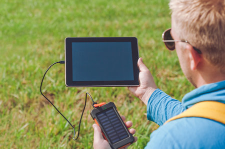 Close-up of a digital tablet in the hands of a man with a charger from a power bank. Lawn green...