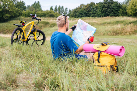 Side view: a man cyclist traveler sits on the green grass and looks at a map in his hands, next to him stands a backpack with a hiking mat. Background of yellow bicycle and nature in the countrysideの写真素材