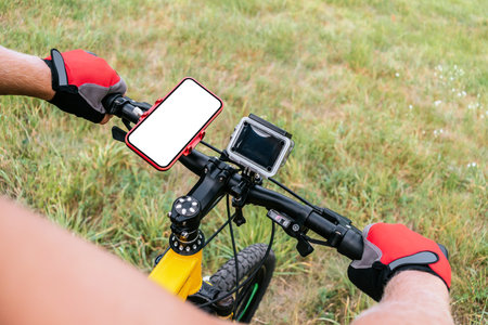 Close-up mockup of a smartphone with a blank white screen on the handlebars of a bicycle with an action camera, man hands in red sports gloves in the countryside on a meadow with green grass. nature background.の写真素材