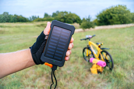 Close-up of using Power Bank charger with solar panel for mobile phone or gadgets, holding in hand of a man in sports cycling gloves, standing on green grass in countryside under blue sky. Background of blurred yellow bicycle and backpack with hiking mat outdoors on a summer sunny day. Battery charge concept, eco friendly technology, portable power source charging smartphone or device while traveling.の写真素材