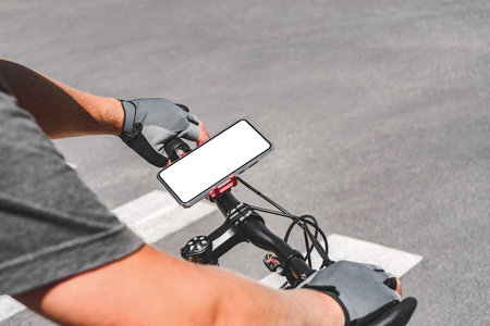 Close-up mockup of smartphone on bicycle handlebar with hands of man in gray cycling gloves. Blank white screen of a mobile phone mounted on bike handlebars. Road background with asphalt or concrete on the roadway.の写真素材