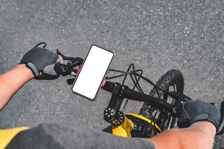 Close-up mockup of a smartphone with a blank white screen of a mobile phone on the handlebar of a bicycle, hands of a man in gray sports cycling gloves. Asphalt road background.の写真素材