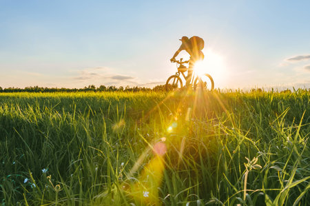Cyclist man tourist with backpack cycling on a bicycle on a field with grass. Background sunset sun with blue sky and silhouette of male bicyclist on bike with rays of sun in the outdoors in the countryside. Side viewの写真素材
