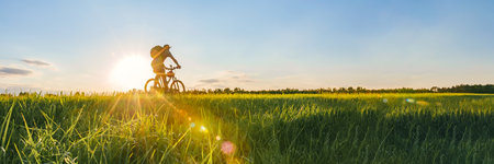 Long Banner, Cyclist riding a bike in a field. Sunset Sun background with blue sky and silhouette of a man bicyclist on a bicycle with backpack and sun rays in the outdoors in the countryside.の写真素材