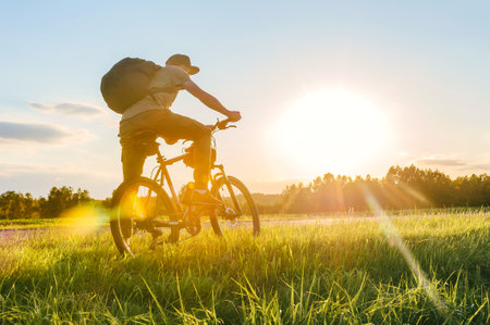 Cyclist riding a bike in a field. Side view of unrecognizable man riding a bicycle on a grassy meadow on a sunny summer evening in the countryside with blue sky at sunset.の写真素材