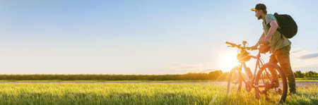 Long Banner, Man cyclist with backpack and cap standing with bicycle in field with green grass and sky. Background sunset sun with traveler on male bike active leisure activity.の写真素材