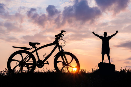 Silhouette of a male cyclist with arms raised above his head, a symbol of freedom with a nearby, standing bicycle. Background sunset sun in the sky. Concept of success, joy of victory and active lifestyle.の写真素材
