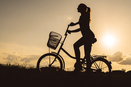 Silhouette of a beautiful unrecognizable girl or woman with a bike in a meadow in a field with grass. Sunset background.の写真素材