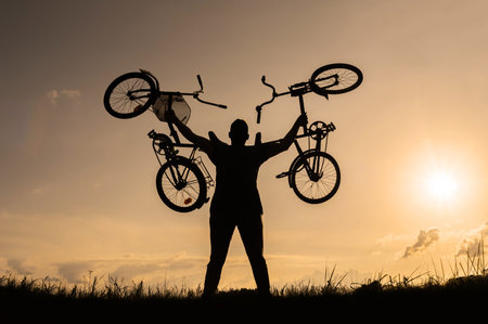 Silhouette of a male cyclist standing in action, lifting bicycles or bicycles above his head. Sky...