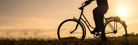 Long Banner. Close-up silhouette of a bicycle with cyclist's legs at sunset. Background of a field in the countryside at dawnの写真素材