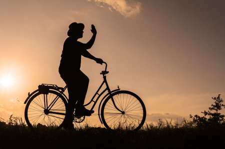 Silhouette of a male cyclist in a hat on a bicycle showing a hi symbol with his hand. Sunset background on a field in the countryside.の写真素材