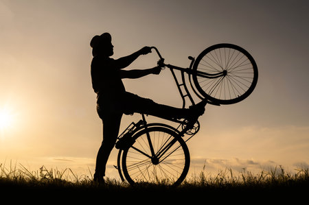 A silhouette of a cyclist holding and standing on the back wheel of a bicycle or bike. Sunset background.の写真素材