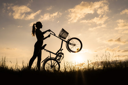 Silhouette of an unrecognizable girl or woman cyclist, standing in a meadow and holding the...