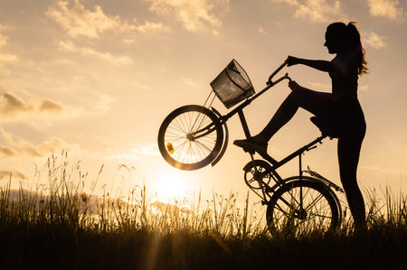 A silhouette of a cyclist woman holding and standing on the back wheel of a bicycle or bike. Sunset backgroundの写真素材